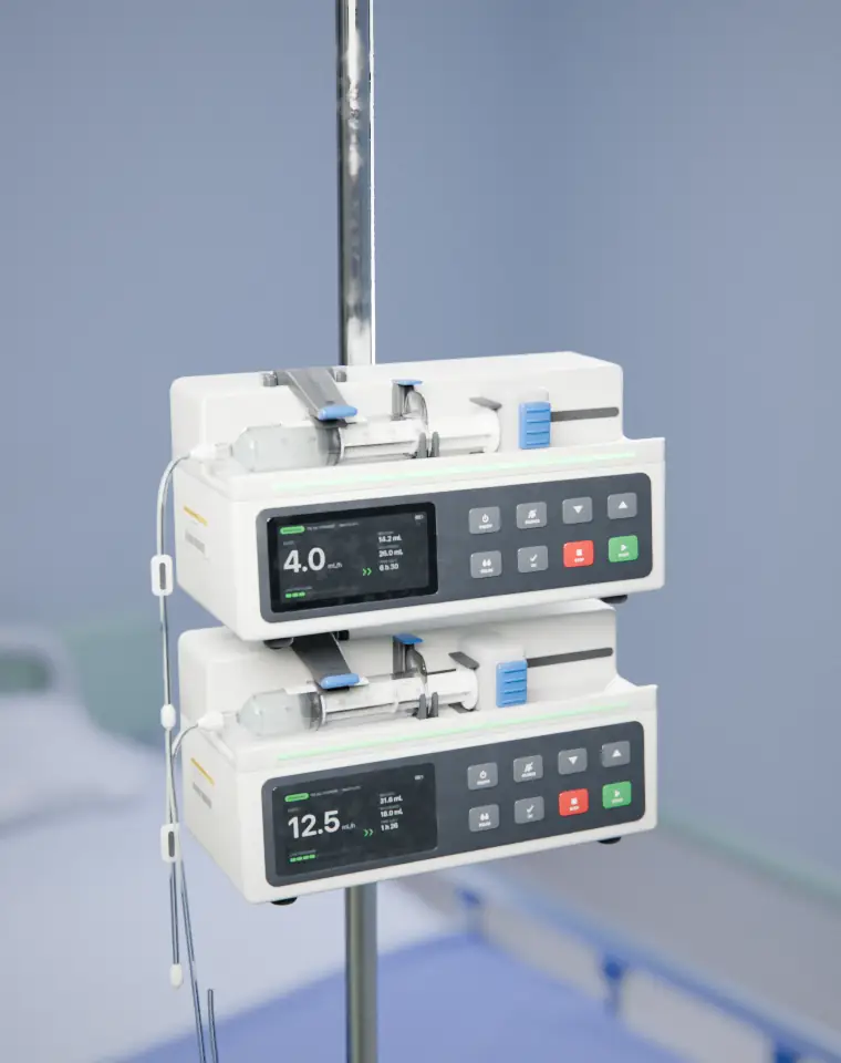 An invented syringe infusion pump in white with a graphite front panel, a lit display and a sample syringe loaded, clamped to an IV pole above a second pump in a hospital ward