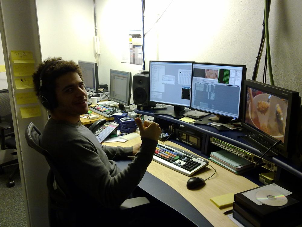 Mo at an edit desk with three monitors, a keyboard and a speaker, giving a thumbs up.