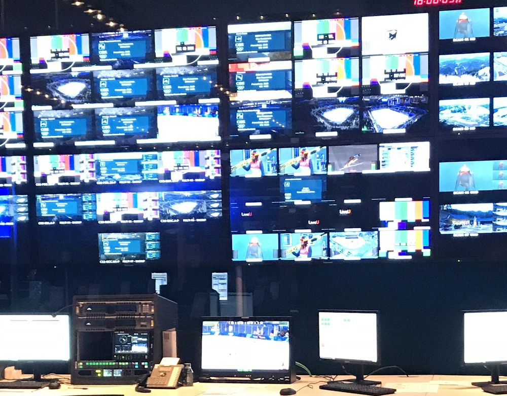 A wall of monitors showing many Olympic feeds above a desk of screens in a broadcast gallery.