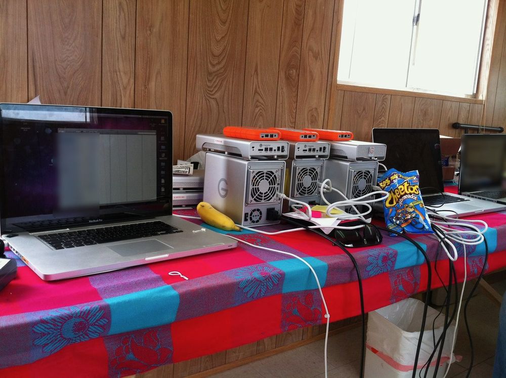 Laptops, a stack of RAID drives and orange rugged drives on a table under a bright cloth.